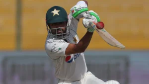 Pakistani cricketer in action during a Test match, wearing a white jersey with a helmet featuring a star emblem, focused on playing a shot with his bat raised.